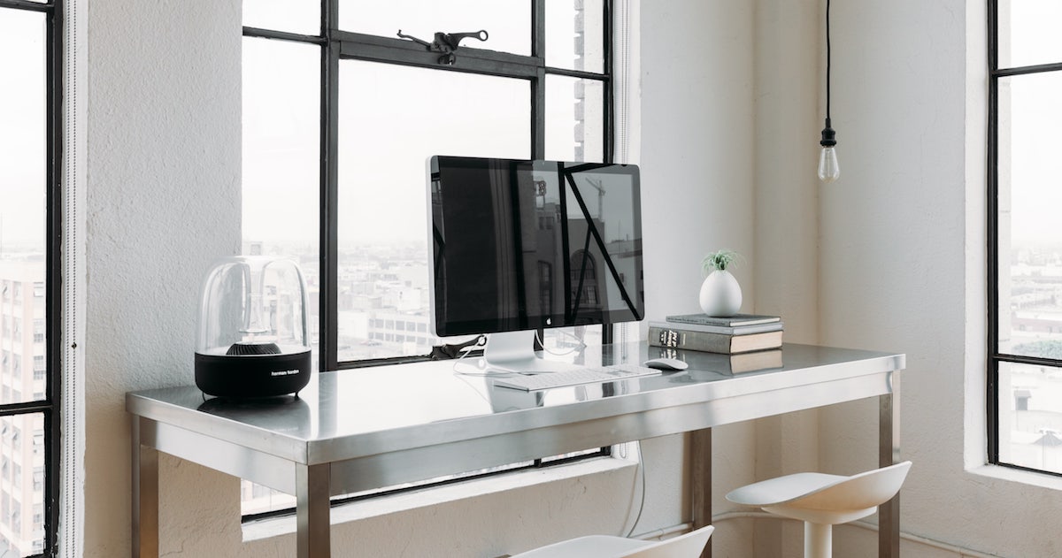Modern desk clear except for streamlined computer sitting against industrial window.